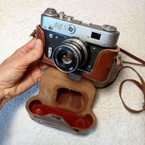 May include: A vintage silver and black FED 3 camera with a lens, in a brown leather case. The camera has a leather strap and is partially out of its case. The case is open, revealing a red interior.