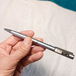 Op de afbeelding: Een zilverkleurige metalen balpen met een klein rechthoekig digitaal LCD-scherm op de houder. Het scherm toont de cijfers 8 25 in zwarte tekst. De pen is voorzien van een zakclip en de cyrillische tekst "ЭЛЕКТРОНИКА 5" op de behuizing.