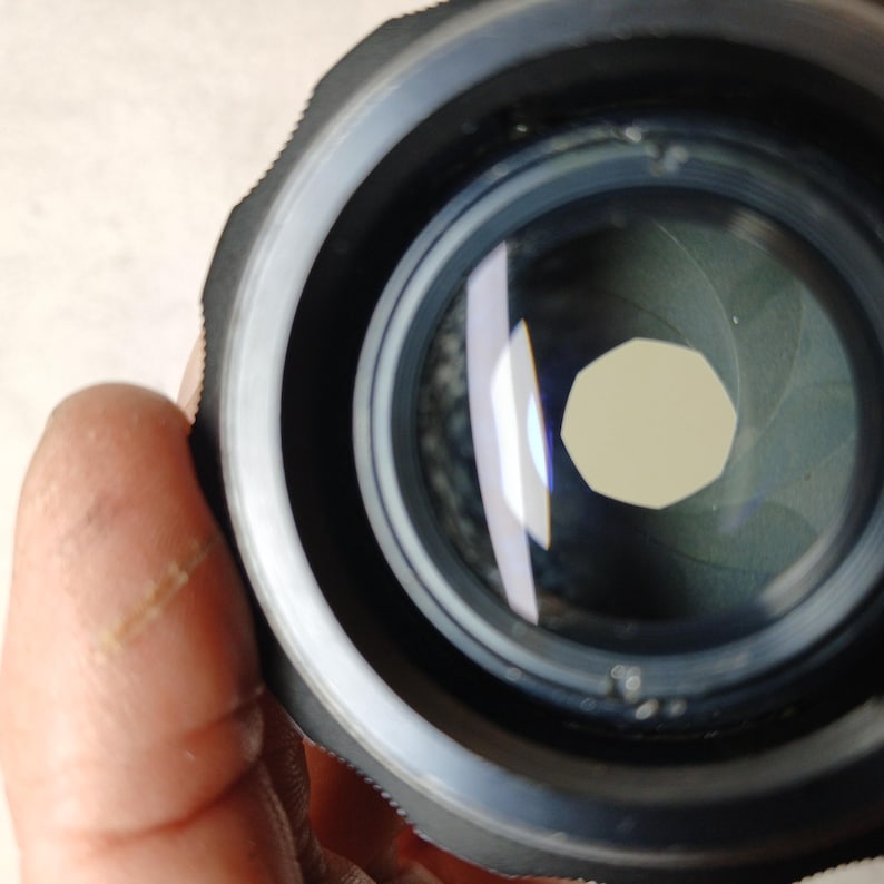 Peut inclure: Gros plan sur un objectif d'appareil photo avec un ext&eacute;rieur noir et argent&eacute;. L'objectif comporte des lames internes formant une ouverture. Le verre est clair, refl&eacute;tant la lumi&egrave;re. L'objectif est tenu par une main.