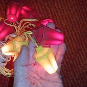 Könnte beinhalten: Eine Lichterkette mit roten und gelben Leuchten. Die Leuchten sind in Form von kleinen Häusern mit einem spitzen Dach. Die Lichter sind an und leuchten.