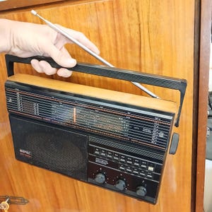 Puede incluir: Una radio portátil con carcasa de madera, panel frontal de plástico negro y asa de transporte negra. La radio cuenta con una antena telescópica plateada larga y fina, una rejilla de altavoz negra, varios botones y tres mandos giratorios.