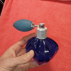 Op de afbeelding: Een vintage kobaltblauwe parfumfles met een geribbeld ontwerp. De fles heeft een zilverkleurige metalen kraag en een blauwe rubberen verstuiverbol. De fles wordt in een hand gehouden tegen een rode achtergrond.