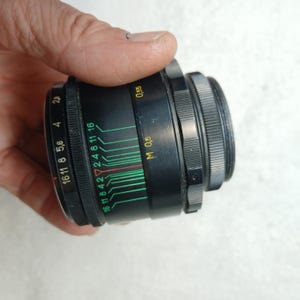 Peut inclure: Un objectif d'appareil photo noir avec une bague de r&eacute;glage argent&eacute;e. L'objectif comporte des marquages verts, rouges et blancs indiquant les r&eacute;glages d'ouverture et de mise au point. L'objectif est tenu dans une main.