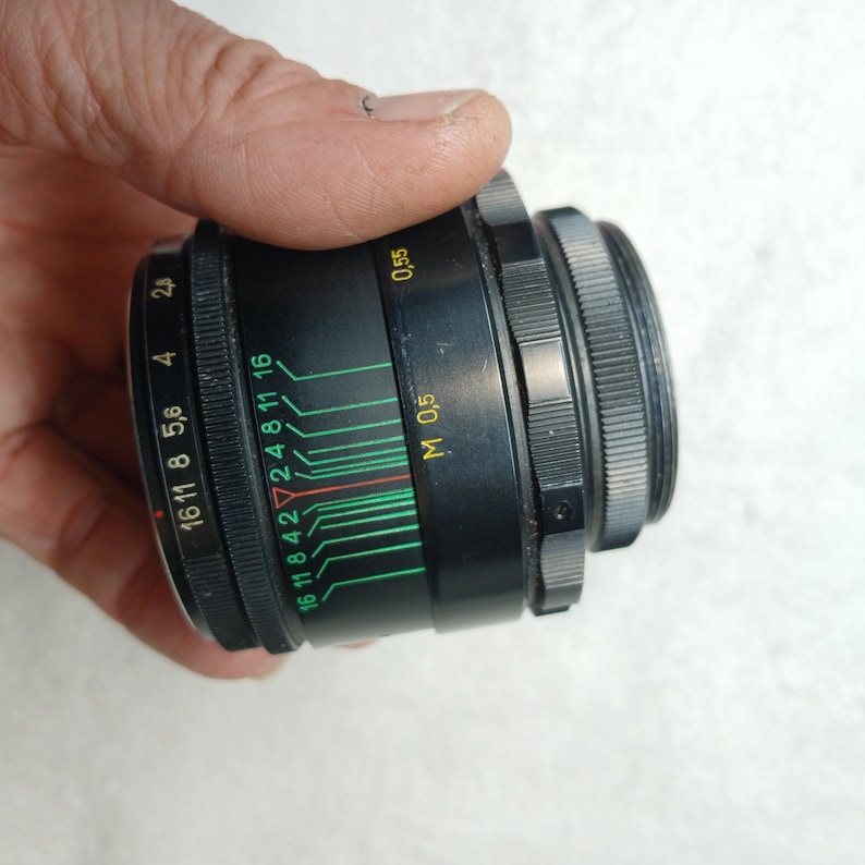 Peut inclure: Un objectif d'appareil photo noir avec une bague de r&eacute;glage argent&eacute;e. L'objectif comporte des marquages verts, rouges et blancs indiquant les r&eacute;glages d'ouverture et de mise au point. L'objectif est tenu dans une main.