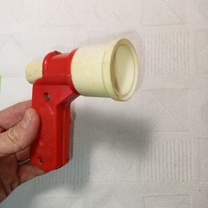 May include: A red and white plastic toy gun with a white barrel and a red handle. The gun has a trigger and a safety.