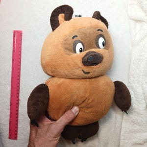 May include: A plush toy shaped like a brown bear with dark brown ears, paws, and a snout. The bear has large white eyes with black pupils and a small stitched smile. A pink ruler is positioned to the left, showing a height of 25 cm.
