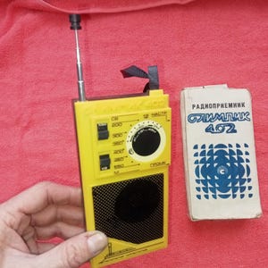 May include: A vintage yellow radio with a retractable antenna and a black speaker grill. The radio has a tuning dial and several buttons. Next to the radio is a white box with blue text that reads "Радиоприемник ОЛИМПИК 402".
