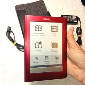 Puede incluir: Un lector electrónico Sony rojo con una pantalla que muestra iconos de menú para leer, tomar notas y configurar. El dispositivo se sostiene en una mano, con un cable de alimentación negro y una funda gris visible. La pantalla muestra el texto "Continue Reading" y "Books".