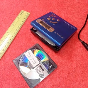 Könnte beinhalten: Ein blauer Sony MiniDisc-Player mit schwarzer Basis, eine MiniDisc und ein gelbes Lineal werden auf einer roten Oberfläche gezeigt. Die MiniDisc befindet sich in einer transparenten Hülle und trägt die Aufschrift "Michael Franks Collection" und "MD 74".