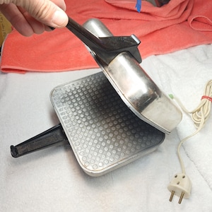 Peut inclure: Un appareil à sandwich électrique argenté avec une surface de cuisson carrée et alvéolée antiadhésive. L'appareil dispose d'un couvercle articulé avec une poignée en plastique noir, une poignée noire assortie sur la base et un cordon blanc.