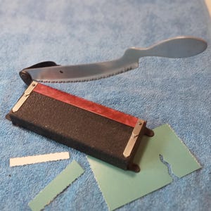 May include: A vintage metal saw with a curved handle and a serrated blade, on a blue cloth. A black and red rectangular tool and several small, cut pieces of paper are also present.