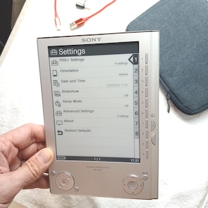 Puede incluir: Un lector electrónico Sony Portable Reader System PRS-505 de color plateado con una pantalla monocromática que muestra un menú de ajustes. El dispositivo cuenta con una pantalla vertical, una barra de navegación lateral numerada y botones circulares.