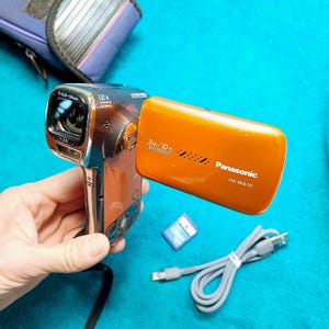 Op de afbeelding: Een oranje Panasonic HX-WA10 waterdichte camcorder met 12x geavanceerde zoom, een waterdichtheid van 3m/10 ft, een blauwe geheugenkaart en een grijze USB-oplaadkabel. Het apparaat heeft een uitklapbaar scherm en een zwarte lensbehuizing.