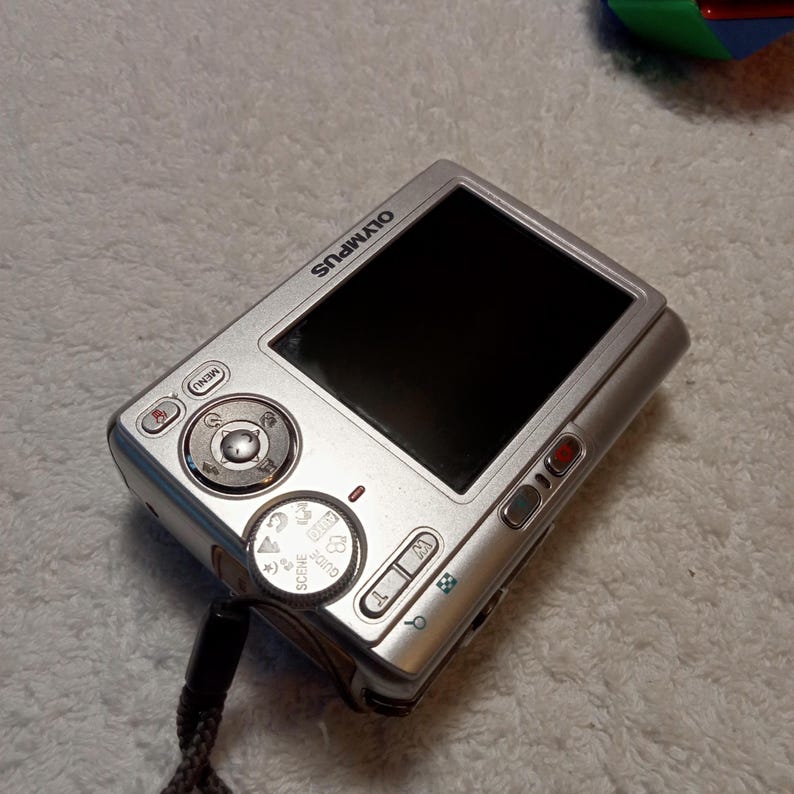 May include: Silver Olympus digital camera with a black screen. The camera has several buttons and dials, including a menu button and a scene dial. A black strap is attached to the camera.