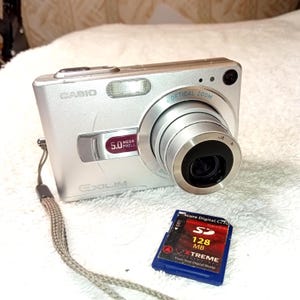 May include: Silver Casio Exilim digital camera with a 5.0 megapixel resolution and optical zoom lens. Includes a blue 128 MB SD card. The camera has a wrist strap attached.