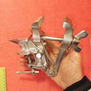 May include: A small, silver-colored bench vise with a red anvil. The vise has a screw mechanism with a butterfly-shaped handle for tightening. The jaws are grooved for gripping. The tool is held in a hand against a red background.