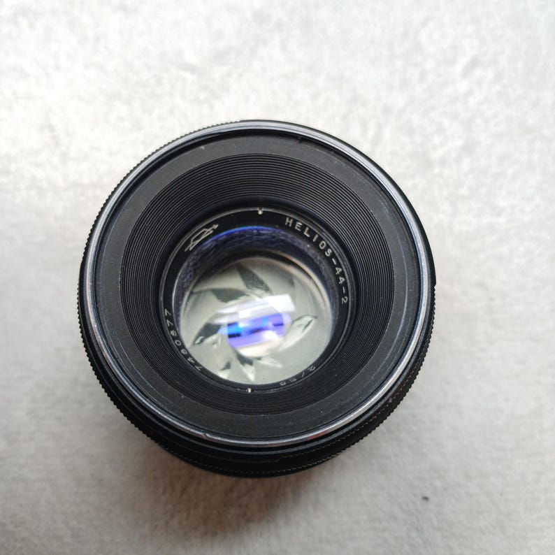 Peut inclure: Un objectif d'appareil photo noir Helios-44-2 avec une bague argent&eacute;e et du texte. L'objectif comporte une s&eacute;rie de lames visibles &agrave; l'int&eacute;rieur, r&eacute;fl&eacute;chissant la lumi&egrave;re. L'objectif est con&ccedil;u pour la photographie.