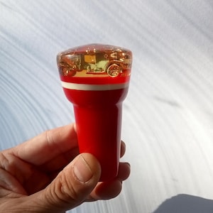May include: A red and white gear shift knob with a clear top. Inside the clear top is a gold-coloured vintage car. The knob is held in a hand.