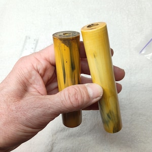 May include: Two cylindrical objects with a yellow and brown color scheme. The objects are held in a hand. The cylinders have a hole in the top and a darker brown color on the bottom. The objects appear to be made of a solid material.