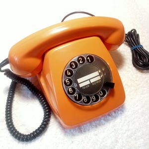May include: Orange vintage rotary dial telephone with a black dial and handset. The phone has a coiled black cord and a label that reads "FEUER NOTRUF". The phone is a retro design.