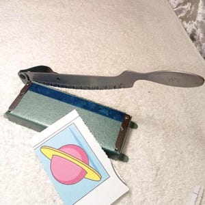 May include: A vintage metal cheese slicer with a serrated blade and a wooden handle. The slicer is paired with a green and blue metal box and a small card featuring a pink planet with a yellow ring.