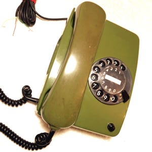 May include: Olive green vintage rotary phone with a black dial and handset. The phone has a coiled cord and a black base. The dial features black numbers on a white background. The phone is a classic design.