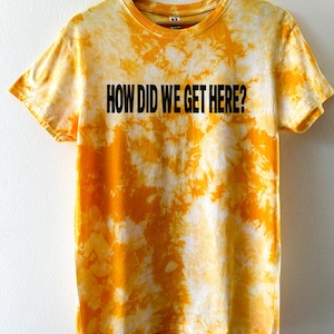 May include: A yellow and white tie-dye t-shirt with the text "HOW DID WE GET HERE?" in black. The t-shirt is displayed on a wooden hanger. The shirt has a crew neck and short sleeves. The background is white.