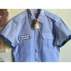 May include: Light blue short-sleeved shirt with two chest pockets and a patch that reads "MR. BLUE COLLAR." The sleeves are trimmed with denim. A small doll with a yellow dress is attached to the shirt with a white string.