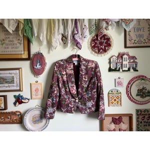 May include: A burgundy blazer with a floral pattern of pink, blue, and green flowers. The blazer is hanging on a pink hanger against a wall decorated with framed art and vintage decor.