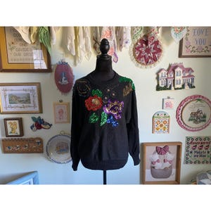 Könnte beinhalten: Ein schwarzer Pullover mit floralen Paillettenverzierungen in Rot, Grün und Lila. Der Pullover wird auf einer Schaufensterpuppe präsentiert. Der Hintergrund umfasst gerahmte Kunstwerke und dekorative Gegenstände.