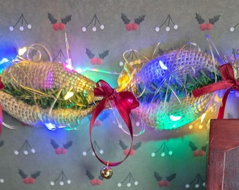 May include: A festive Christmas decoration made of burlap, greenery, and red bows. It is adorned with twinkling multi-colored lights and gold bells.
