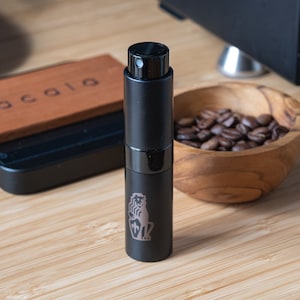 Puede incluir: Una botella de spray negra con una tapa plateada y un pequeño diseño de animal en el lateral. La botella está sobre una superficie de madera junto a un cuenco de granos de café.