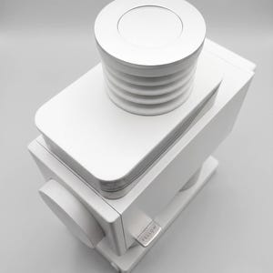 May include: A matte white coffee grinder featuring a rectangular body, a circular dial on the side, and a top-mounted hopper with a ribbed lid. The brand name FELLOW is printed in small, dark letters on a metallic silver tab on the side of the unit.