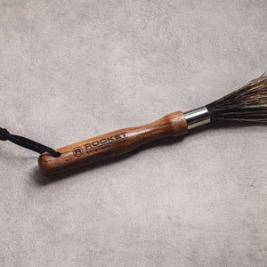 May include: A wooden-handled brush with black bristles and a black leather strap. The handle is engraved with the text "ROCKET" and a logo.