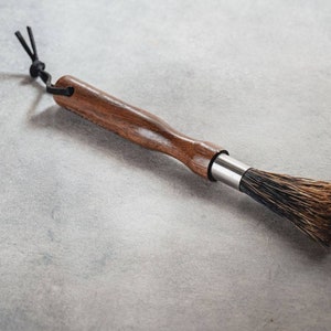 May include: A wooden-handled brush with black bristles and a leather loop. The brush is designed for cleaning coffee grinders.