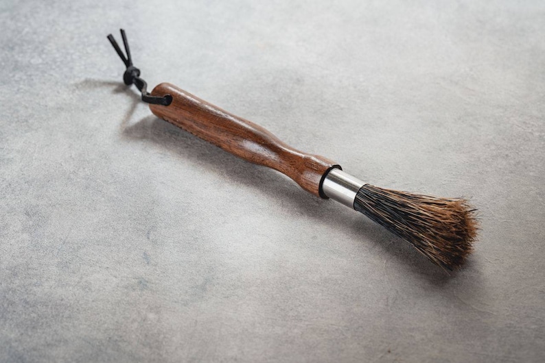 May include: A wooden-handled brush with black bristles and a leather loop. The brush is designed for cleaning coffee grinders.