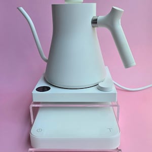 Op de afbeelding: Witte elektrische waterkoker met een zwanenhals-uitloop en een witte basis met een digitaal display en een aan/uit-knop. De waterkoker staat op een transparante acryl standaard.