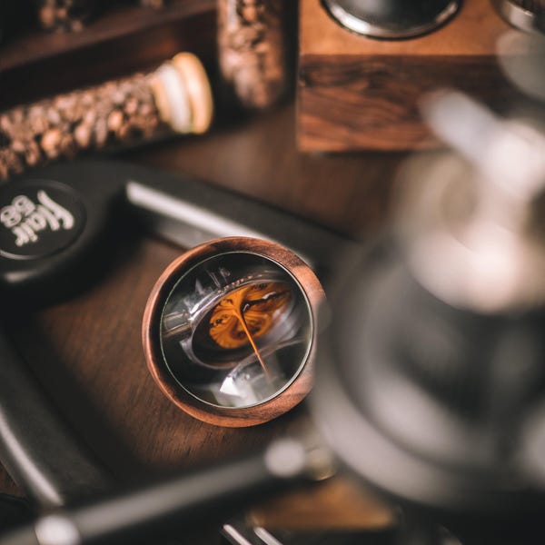May include: A close-up of a coffee machine with a portafilter and a cup of coffee being brewed. The coffee is being poured into a clear glass cup. The coffee machine is made of stainless steel and has a black handle.