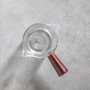 Peut inclure: Une tasse à mesurer en verre transparent avec un corps cylindrique et un bec verseur unique. La tasse présente une poignée en bois brun rougeâtre foncé fixée sur le côté. L'intérieur du verre est lisse et transparent, montrant une base circulaire.