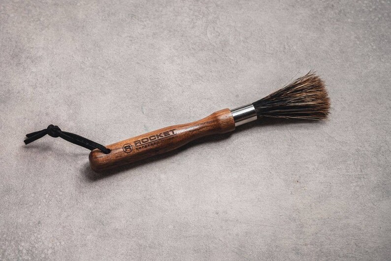 May include: A wooden-handled brush with black bristles and a black leather strap. The handle is engraved with the text "ROCKET" and a logo.