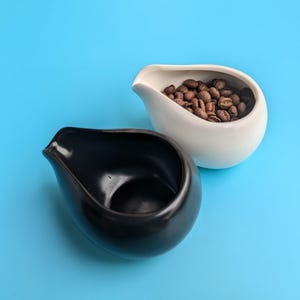 Puede incluir: Dos jarras de vertido de cerámica, una negra y otra blanca, sobre un fondo azul. La jarra blanca contiene granos de café.