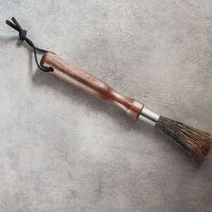 May include: A wooden-handled brush with a black cord loop and natural bristles. The brush is on a gray textured surface.