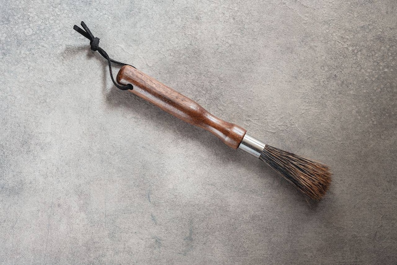 May include: A wooden-handled brush with a black cord loop and natural bristles. The brush is on a gray textured surface.