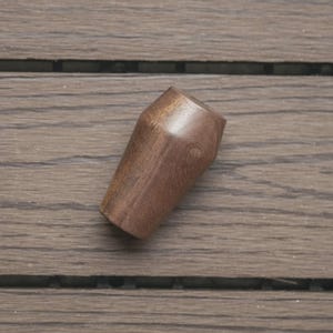 May include: A small, tapered wooden knob with a smooth, polished finish and a rich, dark brown grain pattern. The object features a cylindrical base that widens slightly towards the top, ending in a flat, circular surface at the very peak.