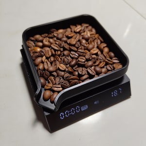 Könnte beinhalten: Eine schwarze digitale Waage mit einem schwarzen Fach, gefüllt mit Kaffeebohnen. Die Waage zeigt "00:00" und "18.7" an.