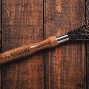 May include: A wooden brush with black bristles and a black leather strap. The brush has the words "ROCKET" and "CARVED WOOD" engraved on the handle.