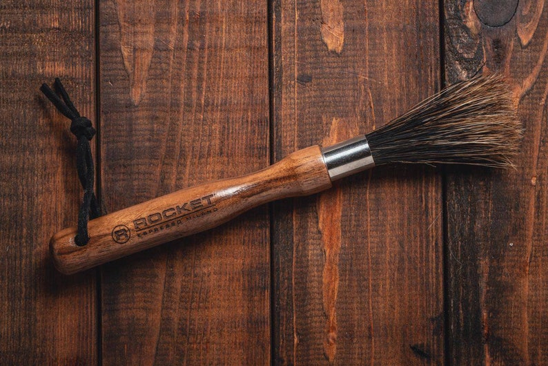 May include: A wooden brush with black bristles and a black leather strap. The brush has the words "ROCKET" and "CARVED WOOD" engraved on the handle.