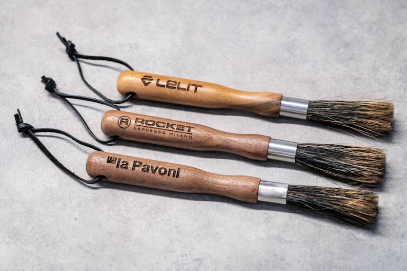 May include: Three wooden-handled cleaning brushes with black leather straps. The brushes are labeled "Lelit", "Rocket Espresso Milano", and "La Pavoni".