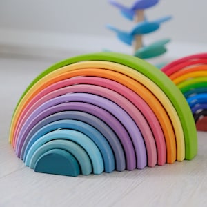 Pode incluir: Um brinquedo de arco de madeira arco-íris com 12 peças. O arco é feito de diferentes tons de verde, amarelo, laranja, rosa, roxo, azul e turquesa.
