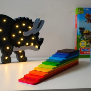 Può includere: Una luce notturna a forma di dinosauro in legno nero con una funzione di illuminazione, un giocattolo di impilamento in legno arcobaleno e un libro blu con il testo "Mein erster Soundbuch" sulla copertina.